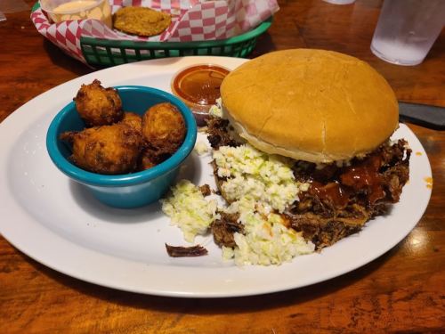 A plate with a large pulled pork sandwich topped with coleslaw, served with a side of fried corn fritters and sauce.