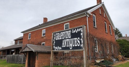 A brick building with a sign reading "Grapevine Antiques" in front, surrounded by greenery and a cloudy sky.
