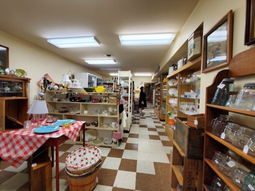 A vintage shop aisle lined with shelves of antiques, dishes, and collectibles, with a checkered tablecloth in the foreground.
