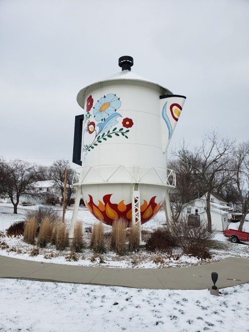A large, decorative water tower shaped like a coffee pot, featuring colorful floral designs and flames at the base, in a snowy landscape.