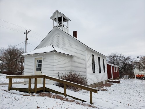 A white schoolhouse with a bell tower, surrounded by snow and a red barn in the background. Overcast sky.