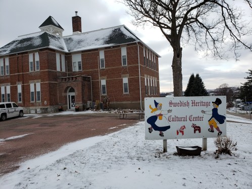 Snow-covered exterior of the Swedish Heritage Cultural Center, featuring a colorful sign and a tree in the foreground.