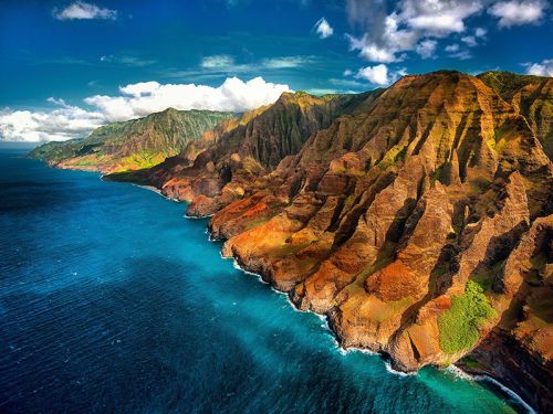 Aerial view of rugged coastal cliffs meeting a vibrant blue ocean under a partly cloudy sky.