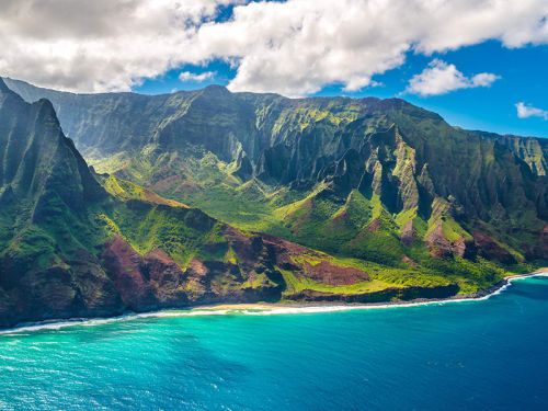 Aerial view of lush green mountains meeting a vibrant blue ocean under a partly cloudy sky.