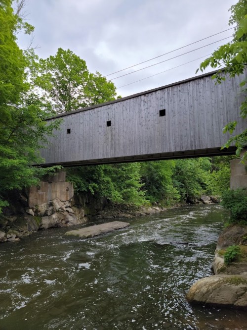 A wooden covered bridge spans a river, surrounded by lush green trees and rocky banks under a cloudy sky.