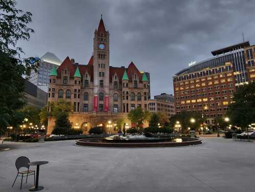 Historic building with a clock tower, surrounded by trees and modern buildings under a cloudy sky.