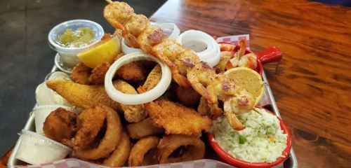 A large platter of assorted fried seafood, onion rings, coleslaw, and a grilled skewer, served with dipping sauces.