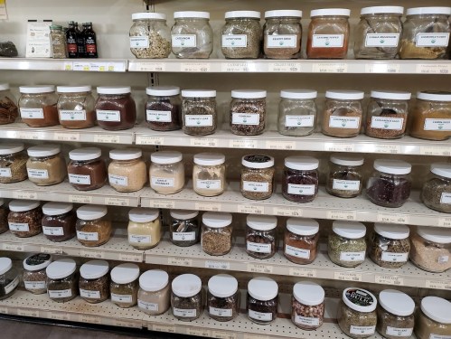 A shelf filled with labeled jars of various grains, seeds, and spices in a store.