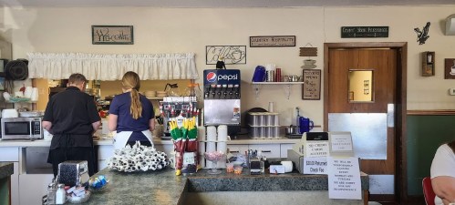 A cozy diner interior with two staff members at the counter, soda fountain, and various condiments on display.