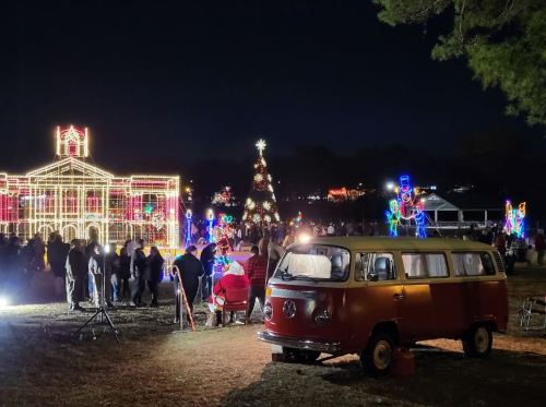 A festive scene with a decorated building, Christmas tree, and a crowd enjoying holiday lights, featuring a vintage van in the foreground.