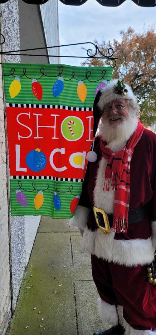 Santa Claus stands next to a colorful "Shop Local" banner decorated with Christmas lights.