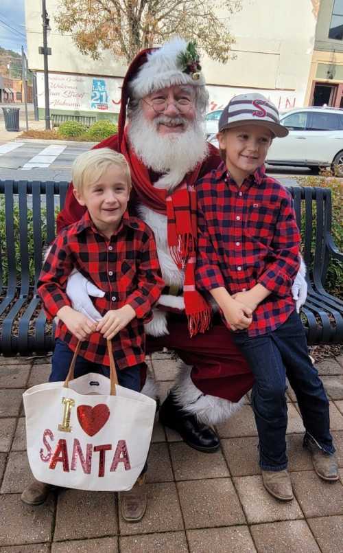 Two children in matching red plaid shirts pose with Santa Claus, who is wearing his traditional outfit and holding a bag.