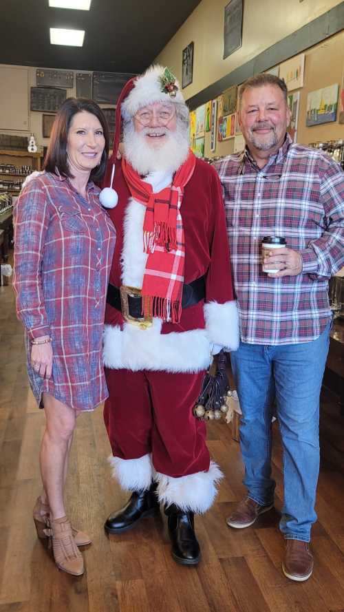 A woman and a man pose with Santa Claus in a festive shop, all smiling and dressed in casual holiday attire.