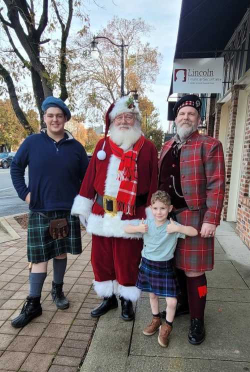 A group of four, including Santa, wearing kilts, posing together on a sidewalk in a festive setting.