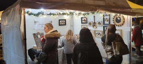 A festive market stall with lights, displaying framed art and crafts, while visitors browse the items.