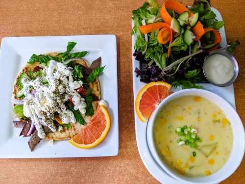 A plate with a flatbread topped with meat, veggies, and sauce, alongside a fresh salad and a bowl of soup.