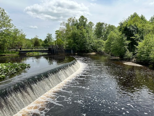 A serene river scene with a waterfall, surrounded by lush green trees and a cloudy sky.