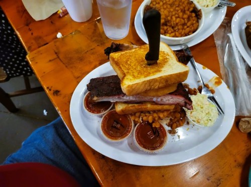A plate of barbecue with stacked toast, brisket, baked beans, coleslaw, and barbecue sauce in cups.