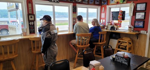 A diner scene with two customers at a window and a staff member in a gray outfit, overlooking a river view.