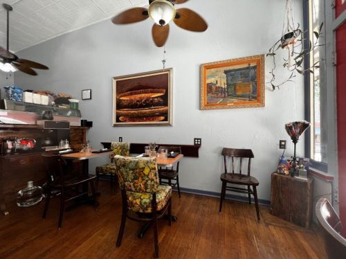 Cozy restaurant interior with wooden tables, floral chairs, and colorful artwork on the walls. Ceiling fans above.