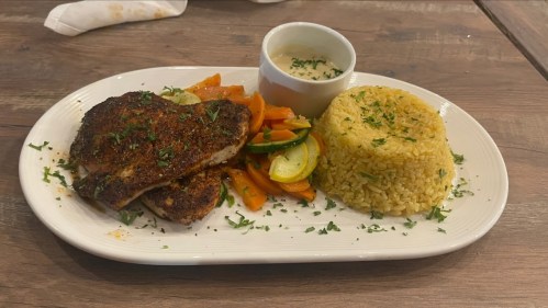 A plate with seasoned grilled fish, sautéed vegetables, yellow rice, and a small bowl of sauce, garnished with herbs.