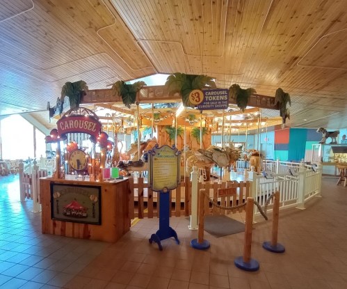 A colorful carousel with animals, surrounded by a wooden fence and signs for tokens, in a spacious indoor area.
