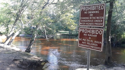 Two signs by a river warning against swimming or standing in the water due to dangerous conditions.