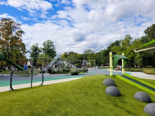 A vibrant playground with green equipment, grassy areas, and trees under a partly cloudy sky.