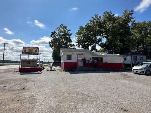 A small, red and white building with a sign, surrounded by gravel and trees, on a sunny day.