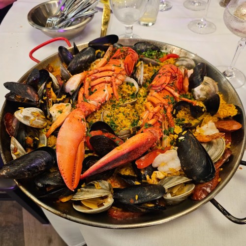 A large paella dish filled with seafood, including lobster, shrimp, mussels, and clams, garnished with herbs.