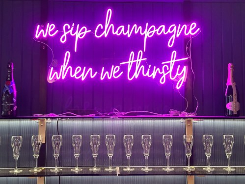 A neon sign reads "we sip champagne when we thirsty" above a row of champagne flutes and bottles.