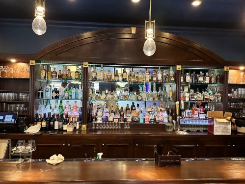 A wooden bar with a variety of liquor bottles displayed on shelves, illuminated by pendant lights.