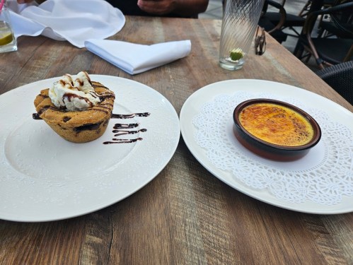 A cookie dessert with ice cream and chocolate drizzle next to a dish of crème brûlée on a wooden table.