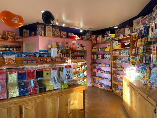 A colorful candy shop filled with shelves of sweets, snacks, and vintage candy displays. Brightly lit and inviting.