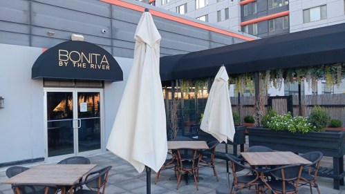 Outdoor seating area of Bonita by the River, featuring tables, chairs, and large umbrellas. Modern building in the background.