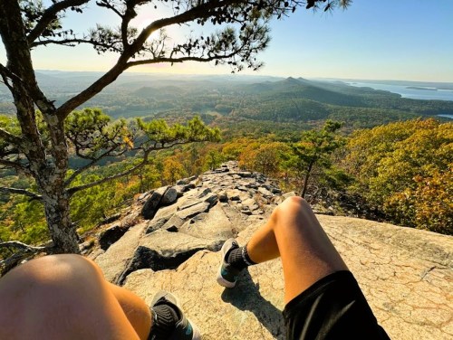 A person sits on a rocky ledge, overlooking a scenic landscape of hills and a lake during sunset.