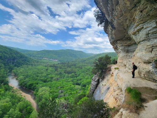 A person stands on a rocky ledge overlooking a lush green valley and river, under a partly cloudy sky.
