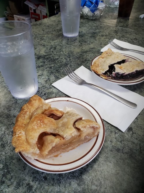 A slice of apple pie on a plate next to a slice of blueberry pie, with glasses of water in the background.