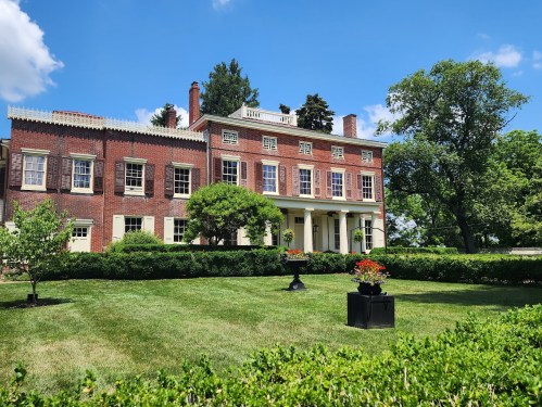 A large brick mansion with white columns, surrounded by green lawns and trees under a blue sky with fluffy clouds.