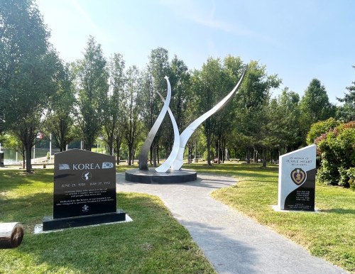 A memorial park featuring a modern sculpture and plaques honoring Korea and Purple Heart recipients, surrounded by trees.
