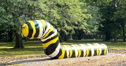 A large, colorful caterpillar sculpture with black and yellow stripes, set in a grassy park area surrounded by trees.