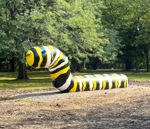 A large, painted caterpillar sculpture in a park, featuring black and yellow stripes, surrounded by trees.