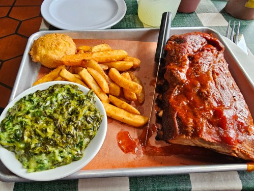 A tray with BBQ ribs, fries, a biscuit, and a side of creamy greens on a checkered tablecloth.