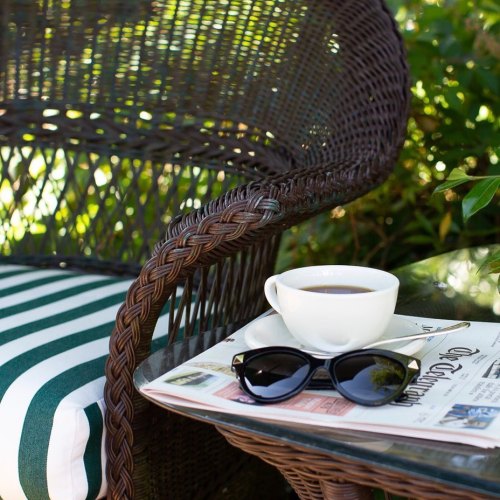 A cozy outdoor scene featuring a wicker chair, a cup of coffee, sunglasses, and a newspaper on a table.