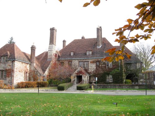 A large, historic stone mansion with a steep roof and multiple chimneys, surrounded by autumn foliage and a manicured lawn.