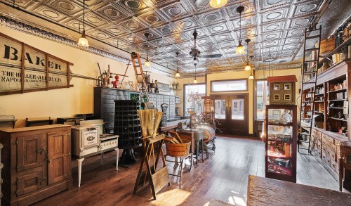 A vintage store interior featuring wooden furniture, antique tools, and a decorative tin ceiling. Warm lighting enhances the atmosphere.