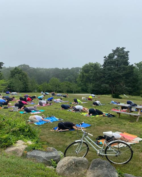 A group of people practicing yoga outdoors on a grassy field, with a bicycle parked nearby and trees in the background.