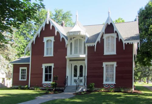 A charming red Victorian-style house with decorative trim, surrounded by greenery and a clear blue sky.