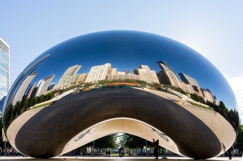 A large, reflective sculpture in a park, mirroring the skyline of a city and surrounding greenery.