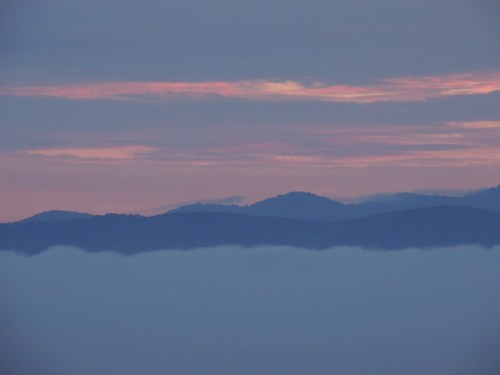 A serene landscape featuring layered mountains beneath a colorful sky at dawn, with clouds blanketing the lower terrain.